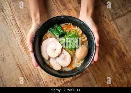 Top view of crop anonymous hands of person holding bowl of ramen soup with pork meat herbs vegetables and boiled egg on wooden table Stock Photo