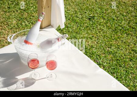 From above of pair of glasses with rose champagne placed near bowl with bottles and ice on canopy bed placed on green meadow during romantic celebrati Stock Photo