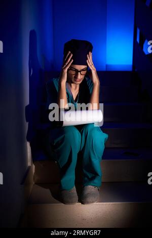 Full body of young thoughtful female doctor in uniform with stethoscope sitting on staircase in room with neon light and touching head while reading d Stock Photo