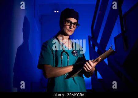 Professional female doctor in medical uniform with stethoscope writing on clipboard and looking at camera while standing on stairs in dark room under Stock Photo