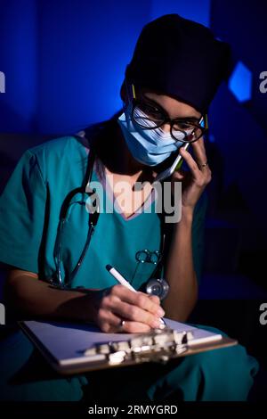Concentrated female doctor in uniform and mask sitting on stairs with clipboard and making notes in clinical record while speaking on smartphone in co Stock Photo