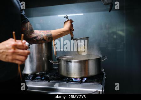 Side view of crop anonymous tattooed male cook standing near plate and preparing noodles in pot in kitchen Stock Photo