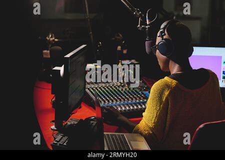 Back view of unrecognizable ethnic female in headphones using mixing console while sitting at table with laptop in dark broadcast studio and talking i Stock Photo