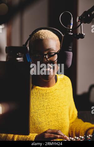 Smiling ethnic female radio host in vivid yellow sweater working in broadcast studio while using mixing console and speaking in microphone Stock Photo