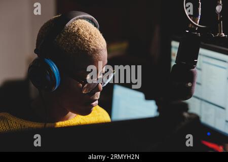 Professional African American female radio host in headphones sitting at table with monitors and looking down while working in modern broadcast studio Stock Photo