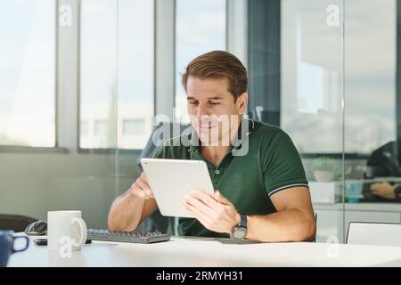 Professional male entrepreneur in green polo sitting at desk with keyboard and browsing tablet while working on project in creative workspace Stock Photo
