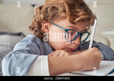 Concentrated preteen boy in casual clothes and eyeglasses sitting at desk while looking down and taking notes while doing homework Stock Photo
