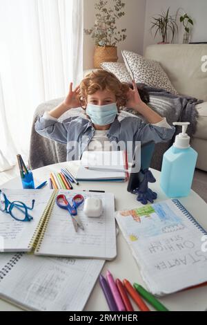 High angle of boy sitting at table with school supplies wearing protective mask in living room during coronavirus outbreak at home Stock Photo