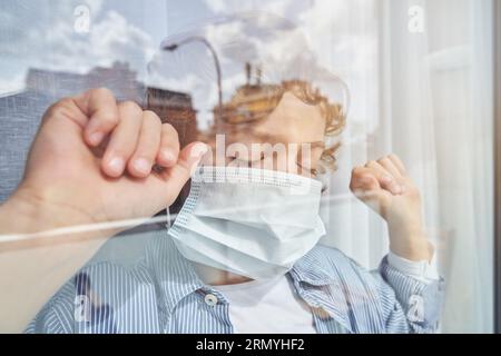 Through glass view of boy with curly blond hair in casual clothes and medical mask standing near window with closed eyes and clenched fists during COV Stock Photo