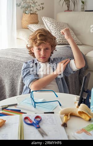 Displeased preteen boy showing muscles and frowning while sitting at table with medical mask and stationery and doing homework in living room at home Stock Photo