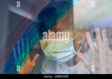 Preteen kid with curly hair wearing protective face mask standing with closed eyes near window with colorful rainbow during coronavirus epidemic Stock Photo