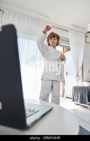 Low angle of concentrated child in white kimono watching video on laptop and doing upward block during training in light living room at home Stock Photo