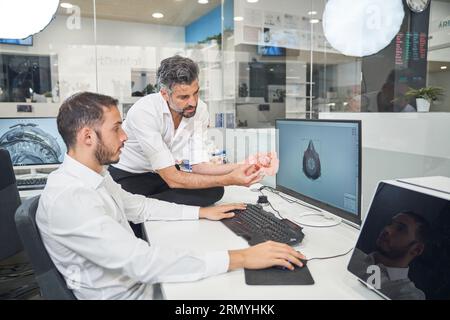 Professional male engineers examining 3D printed respirator while working at table on computer with draft of face mask in office during coronavirus pa Stock Photo