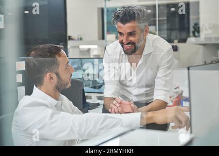Smiling bearded male engineers in white shirts looking at each other while working on computer together in modern light office Stock Photo