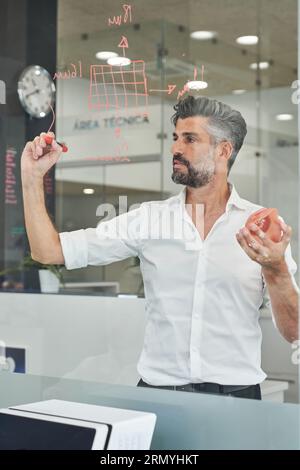 Trough window of serious male engineer in formal wear taking notes on glass wall while designing model of face mask Stock Photo