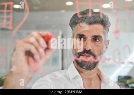 Through glass view of unshaven Hispanic man in shirt writing information on glass wall and working at office Stock Photo