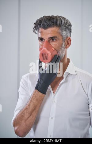 Serious bearded male wearing respirator and medical gloves touching chin and preventing infection spread while standing on white background Stock Photo