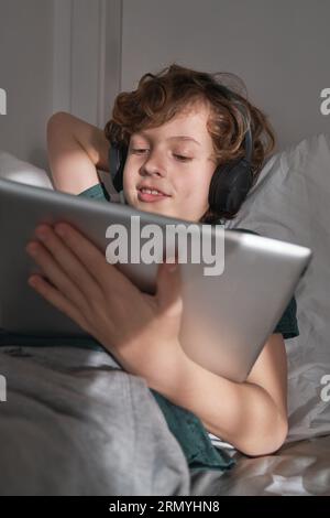 From below of cheerful preteen curly haired boy lying on soft pillow with tablet and watching video in headphones Stock Photo