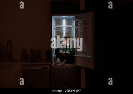 Full body of pensive kid in domestic clothes sitting in fridge with tablet and browsing internet in dark kitchen at night Stock Photo