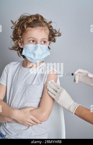 Crop nurse in gloves making injection with COVID 19 vaccine to curly haired child in medical mask against white wall in clinic Stock Photo