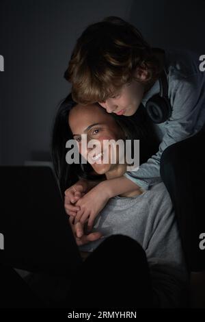 Cheerful curly haired preteen boy with headphones hugging smiling mother while watching film on laptop together in dark room Stock Photo