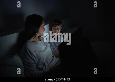 Smiling woman and boy in sleepwear lying on bed with laptop while watching movie at night and looking at each other Stock Photo