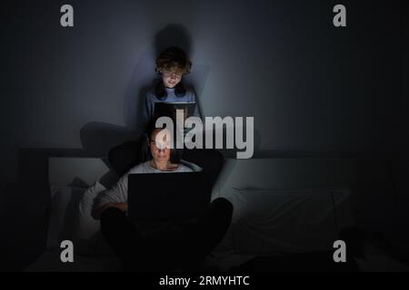 Concentrated woman lying on bed and working on laptop near curly haired boy sitting at headboard and surfing internet on tablet at night Stock Photo
