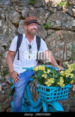 Cheerful male in casual clothes smiling and looking away while sitting on blue flowerbed in form of bicycle with blooming yellow flowers against old s Stock Photo