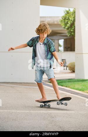 Full body of concentrated child in casual clothes looking down while learning to ride skateboard on city street on bright summer day Stock Photo