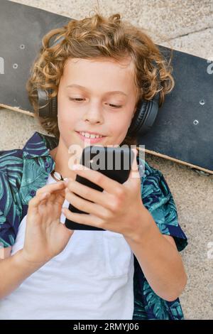 Top view of smiling boy listening to music in headphones and surfing social media on mobile phone while lying on skateboard on city street Stock Photo