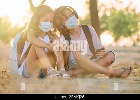 Full length of brother and sister in protective masks listening to music using headphones and sitting on ground in woods during COVID pandemic Stock Photo