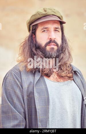 Portrait of serious bearded adult male with long wavy hair and mustache in unusual outfit and cap Stock Photo