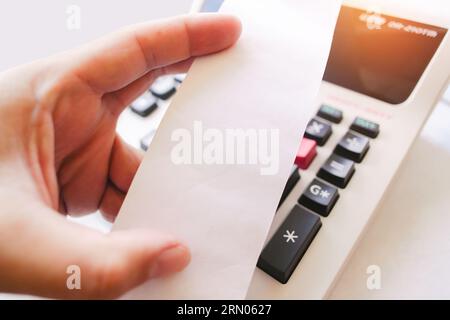 Hand holding blank white paper slip with printing calculator on table, Close up a white paper slip of printing calculator, The concept of financial op Stock Photo