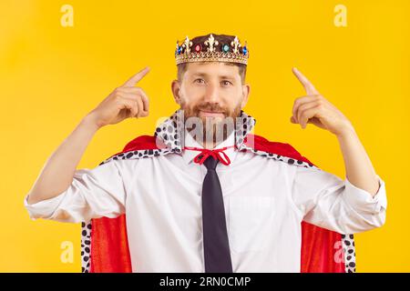 Portrait of young confident man in suit with crown looking at camera on yellow background. Stock Photo