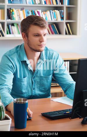 Young man sits at table in room with books and looks into computer monitor. Independent distance learning. Caucasian guy is studying at home. Stock Photo