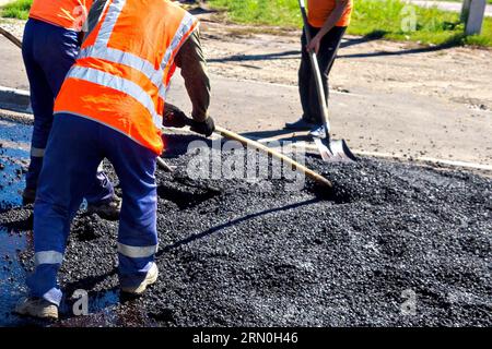 Road workers laying asphalt with shovels on street on summer day. Repair and restoration of road surface. Stock Photo