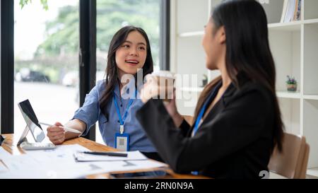 Two happy and charming Asian businesswomen or office workers are enjoying talking during a coffee break at a table in the office together. Stock Photo