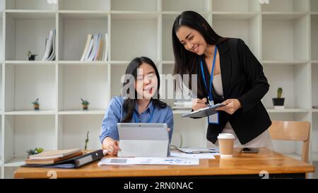 Two pretty Asian businesswomen are working on a project in the office together. colleagues co-working, sharing ideas, brainstorming Stock Photo