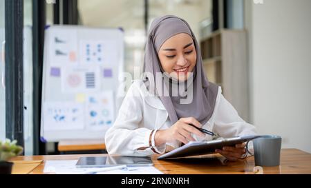 A beautiful Asian Muslim businesswoman wearing a hijab is reading documents, examining business report at her desk. Stock Photo
