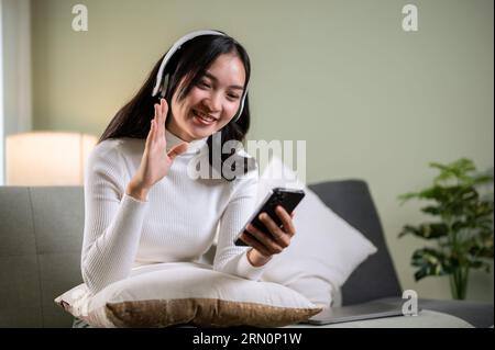 A cheerful and beautiful young Asian woman wearing headphones is enjoying chatting with her friend on a video call through her smartphone while relaxi Stock Photo