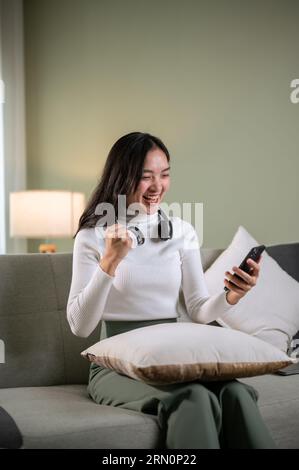 A joyful and overjoyed Asian woman in comfy clothes receives good news on the phone, shows her fist, and screams with happiness and excitement while s Stock Photo