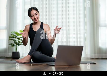 An attractive and sporty Asian female online yoga teacher or trainer is training her students online at home or coaching a virtual yoga class on a vid Stock Photo