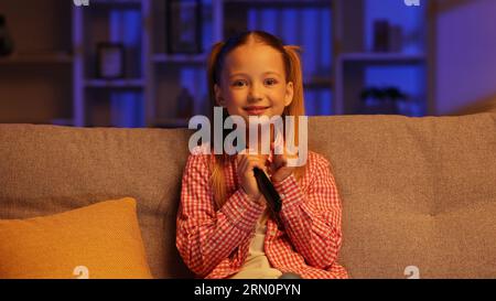 Cheerful girl is sitting on the sofa in the evening and turns on the TV with the remote control. Girl is happy that her favorite cartoon program is on Stock Photo