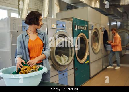 smiling young asian woman looking at boyfriend near clothes in basin in blurred coin laundry Stock Photo