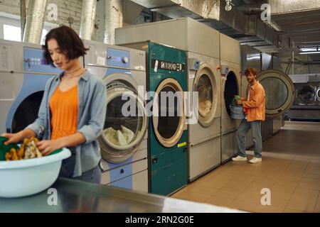 young man standing near washing machine and blurred girlfriend with clothes in coin laundry Stock Photo