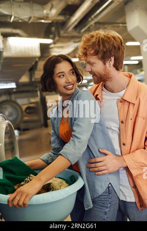 redhead man hugging cheerful asian girlfriend with clothes in basin in public laundry Stock Photo
