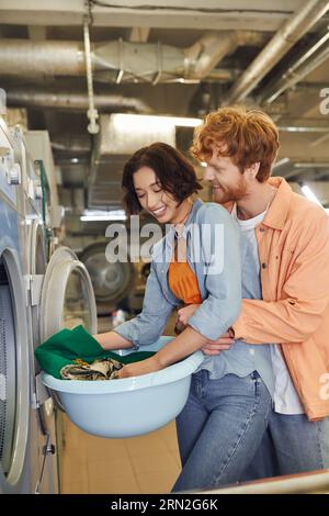 smiling young man hugging asian girlfriend with clothes near washing machine in public laundry Stock Photo