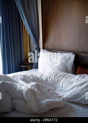Crumpled white bedding sheets linen with pillow and blanket in soft morning lights, comfortable mood of rest and relax bedroom, unmade empty bed hotel Stock Photo