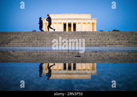 WASHINGTON, DC, United States — Two joggers run past the Lincoln Memorial in the predawn hours, their silhouettes and the iconic monument reflected in a puddle on the plaza steps. The scene captures the interplay between the city's daily life and its historic landmarks, as early morning exercise enthusiasts share the space with one of America's most revered monuments before the day's tourists arrive. Stock Photo