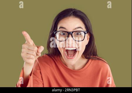Happy amazed woman in glasses pointing finger in camera in orange sweatshirt on khaki background. Stock Photo